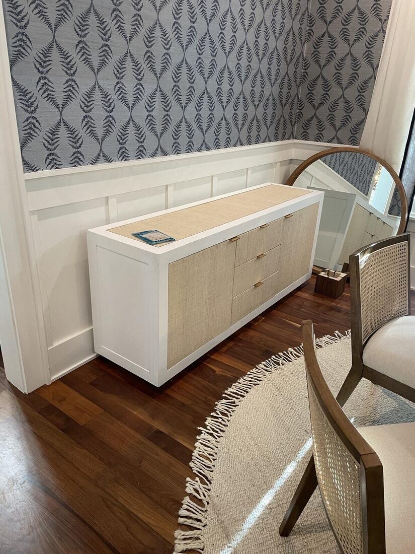 Custom white built-in cabinet with woven rattan door and drawer fronts, set against wainscoting and a round mirror.